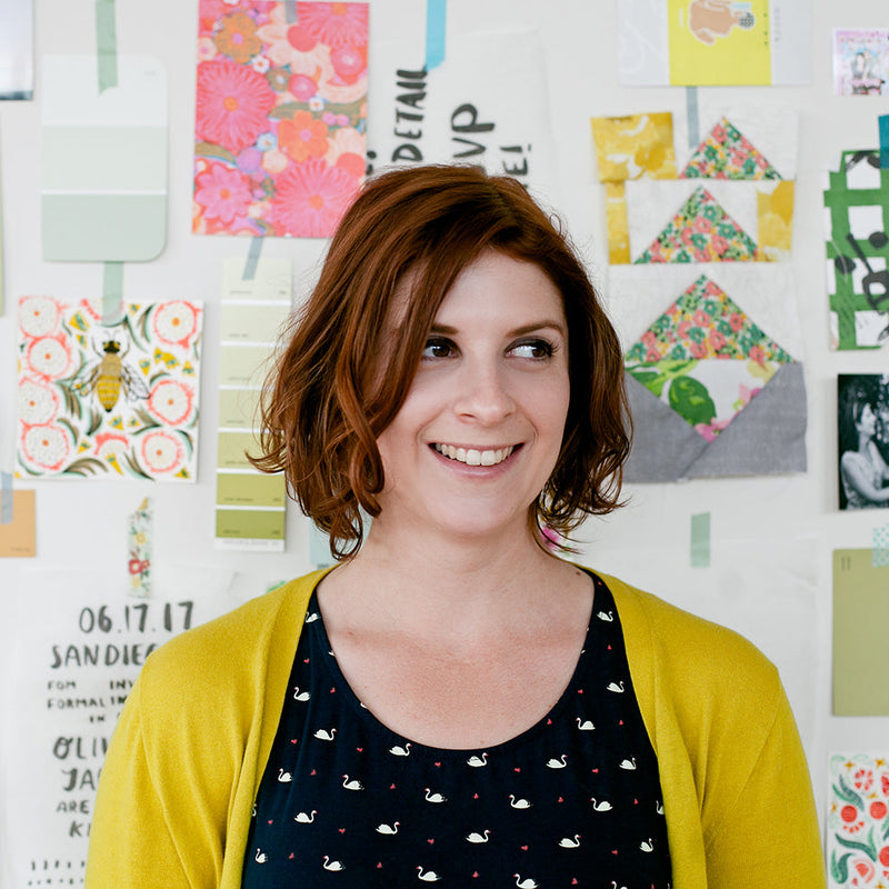 A smiling woman with short auburn hair stands in front of a white wall covered with colorful printed cards, swatches, and small artwork. She is wearing a mustard yellow cardigan over a black top with a small white pattern.